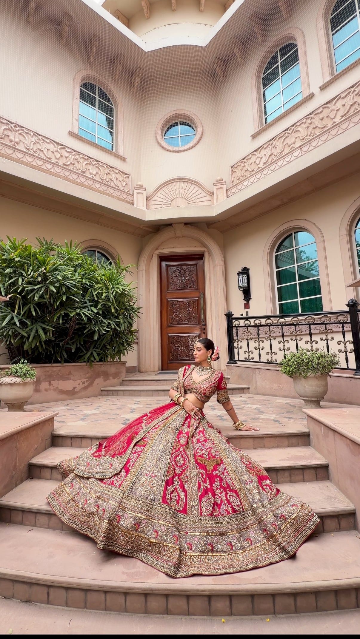 Bride in a red and gold lehenga with soft-glam bridal makeup by Kamya Mehta, photographed on the steps of a heritage venue